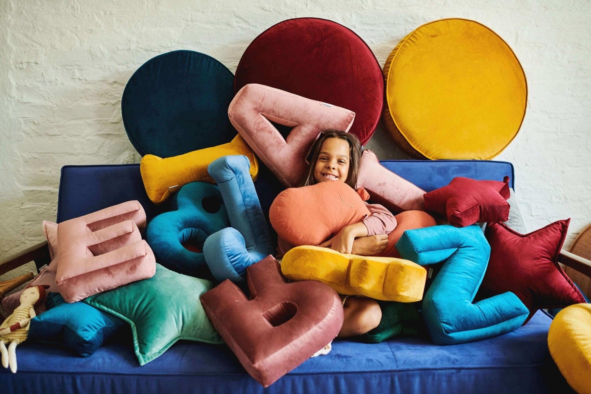 girl sitting on sofa surrounded by many letter cushions and a large velour pouffe from bettys home which are gifts for twin